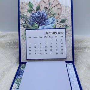 May include: A blue and white desk calendar for January 2026, featuring a floral design and a clock image. The calendar is open, revealing a notepad. The calendar is approximately 6 inches tall.