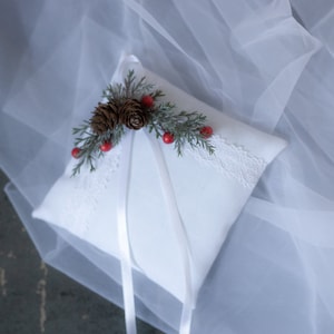 May include: White ring pillow with a decorative arrangement of pinecones, greenery, and red berries. The pillow has a lace trim and white satin ribbons. A wedding accessory.