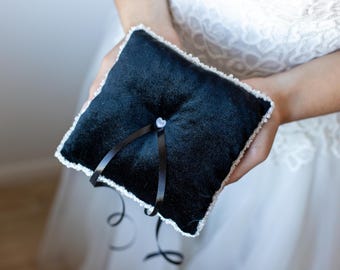 May include: A black velvet ring pillow with a white lace trim and a black satin ribbon. The pillow is held in the hands of a person.