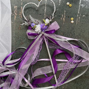 May include: A decorative wand with a heart-shaped top, adorned with purple and white flowers, silver accents, and flowing ribbons. The wand features a silver handle and is embellished with a bow and lace details. The ribbons are in shades of purple and silver.