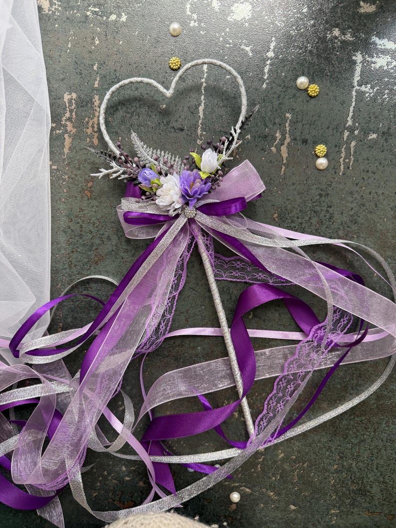 May include: A decorative wand with a heart-shaped top, adorned with purple and white flowers, silver accents, and flowing ribbons. The wand features a silver handle and is embellished with a bow and lace details. The ribbons are in shades of purple and silver.
