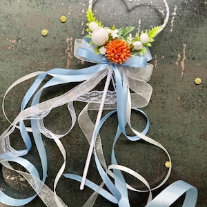 May include: A decorative wand with a heart-shaped top, adorned with white and orange flowers, green leaves, and a light blue bow. Long, flowing ribbons in white and light blue cascade from the base, creating a whimsical and elegant design. The wand is perfect for weddings.