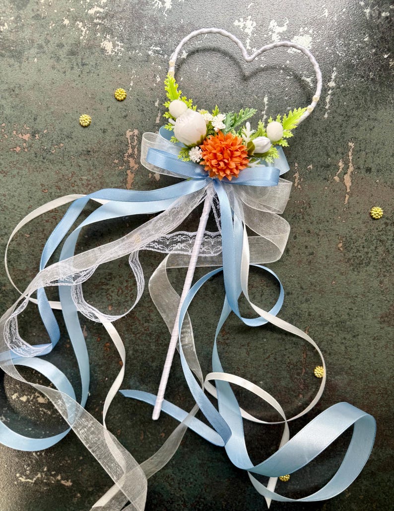 May include: A decorative wand with a heart-shaped top, adorned with white and orange flowers, green leaves, and a light blue bow. Long, flowing ribbons in white and light blue cascade from the base, creating a whimsical and elegant design. The wand is perfect for weddings.