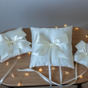 May include: Three ivory satin ring bearer pillows with large satin bows. The pillows are arranged on a wooden table, with a string of star-shaped fairy lights. The pillows are different sizes.