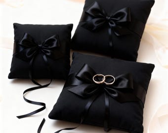 May include: Three black ring bearer pillows with satin bows. Two gold wedding rings rest on the center pillow. The pillows are square and have a ribbon tied around them with a bow.