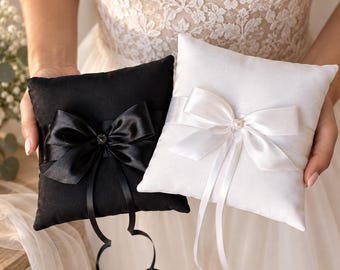 May include: Two square ring bearer pillows, one in solid black and one in solid white, both crafted from smooth fabric. Each pillow features a wide satin ribbon band wrapped horizontally, centered with a decorative bow and a small crystal accent.