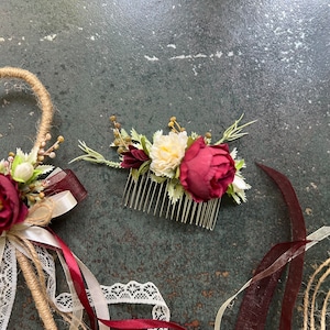 May include: A floral hair comb with a burgundy peony and cream-colored flowers, accented with greenery. The comb is made of gold-colored metal. Ribbons and a decorative wand with flowers and lace are also visible.