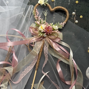 May include: A heart-shaped wedding wand with a wooden handle, wrapped in twine. The heart is adorned with small flowers and greenery. Ribbons in shades of pink, cream, and gold cascade from the wand, creating a whimsical and romantic aesthetic.