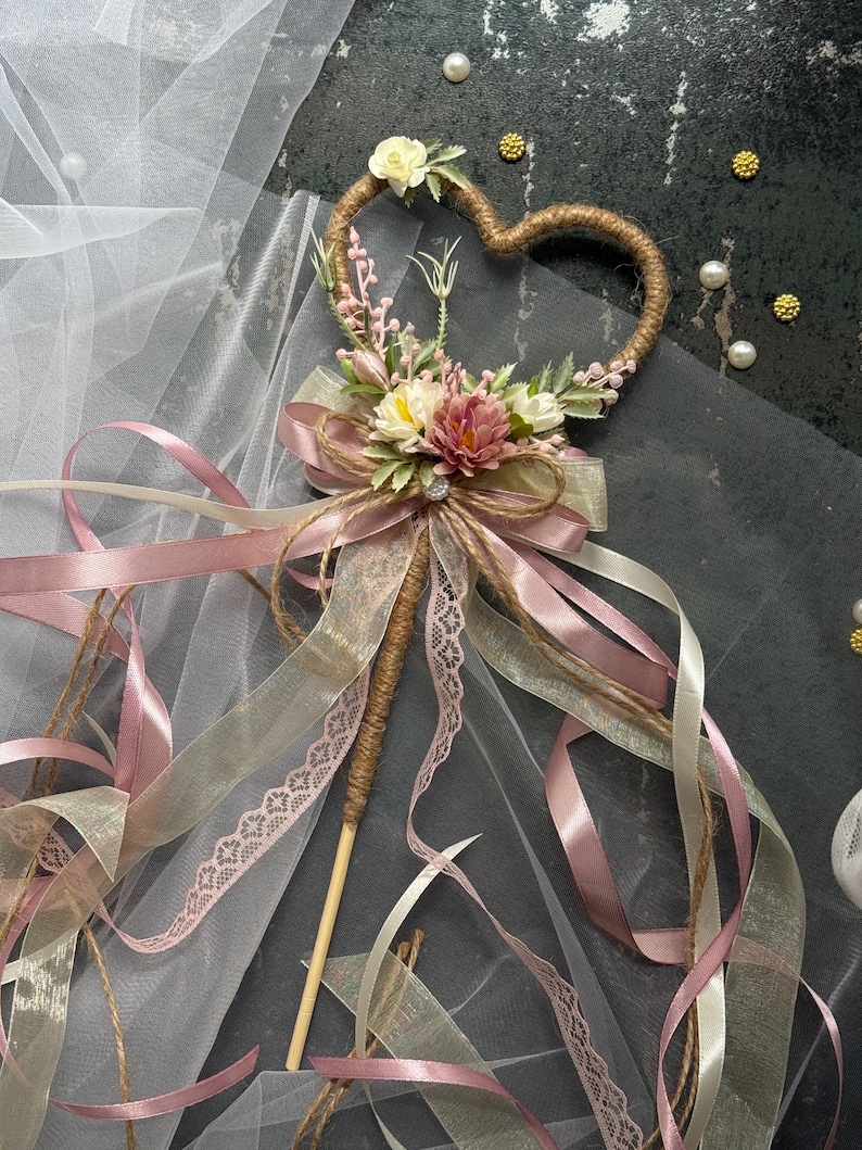 May include: A heart-shaped wedding wand with a wooden handle, wrapped in twine. The heart is adorned with small flowers and greenery. Ribbons in shades of pink, cream, and gold cascade from the wand, creating a whimsical and romantic aesthetic.