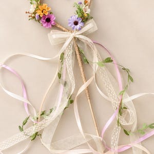 May include: A heart-shaped wand wrapped in twine, adorned with small purple, orange, and white artificial flowers. A sheer cream ribbon is tied in a bow at the base of the heart, with long trailing ribbons in cream and light pink, plus green vines.