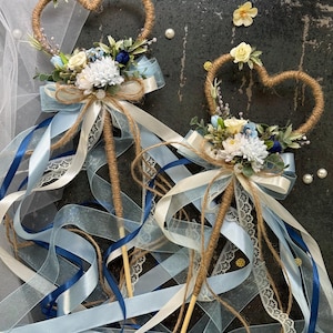 May include: Two heart-shaped wedding wands. The wands have a twine heart, floral arrangement, and ribbons in shades of blue and white. The wands are decorated with white lace and pearls. The wands are perfect for a wedding.