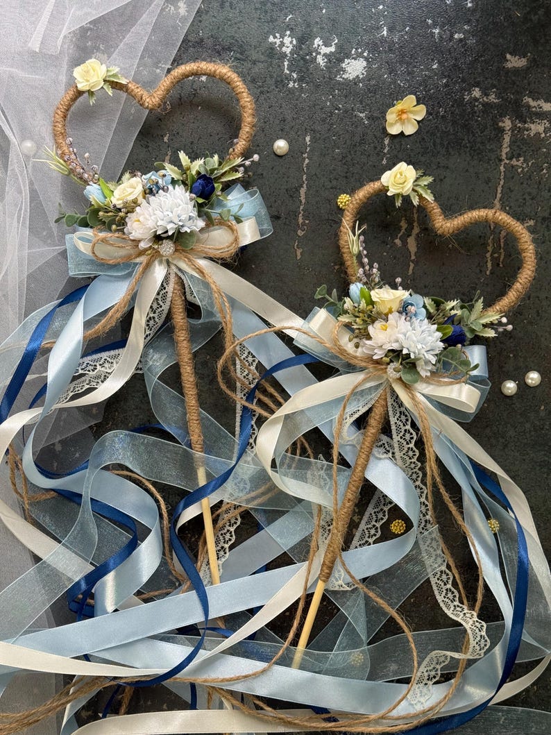 May include: Two heart-shaped wedding wands. The wands have a twine heart, floral arrangement, and ribbons in shades of blue and white. The wands are decorated with white lace and pearls. The wands are perfect for a wedding.