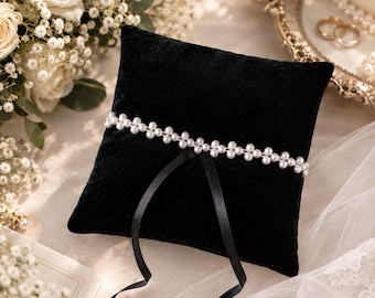 May include: A square black velvet ring pillow featuring a horizontal band of small white faux pearls across the center. Two thin black satin ribbons are attached to the pillow, extending downward from the pearl band to secure rings.