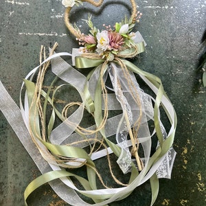 May include: A heart-shaped wedding wand with a twine frame, adorned with white and pink flowers, green leaves, and a green satin bow. White and silver ribbons and lace cascade from the base, creating a whimsical and elegant design.