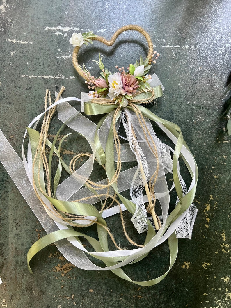 May include: A heart-shaped wedding wand with a twine frame, adorned with white and pink flowers, green leaves, and a green satin bow. White and silver ribbons and lace cascade from the base, creating a whimsical and elegant design.