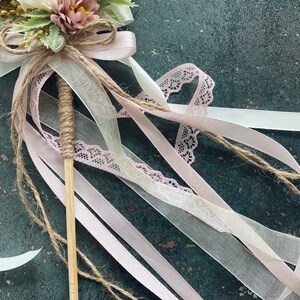 May include: A decorative wand featuring a bouquet of white and mauve flowers, accented with greenery and tied with a jute bow. Pink and white ribbons, along with a lace ribbon, cascade from the floral arrangement. The wand's handle is wrapped in twine.