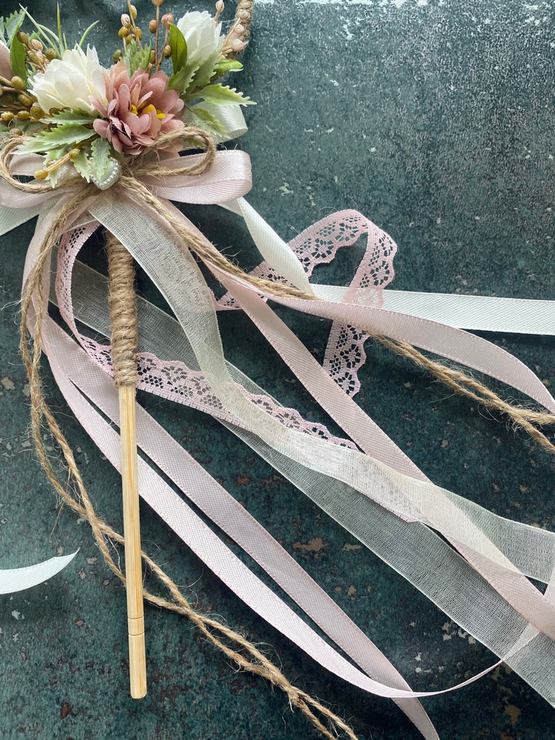 May include: A decorative wand featuring a bouquet of white and mauve flowers, accented with greenery and tied with a jute bow. Pink and white ribbons, along with a lace ribbon, cascade from the floral arrangement. The wand's handle is wrapped in twine.