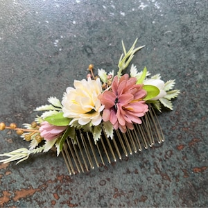 May include: A floral hair comb featuring a cluster of artificial flowers in shades of cream, pink, and mauve, accented with green leaves and sprigs. The flowers are attached to a gold-colored metal comb, suitable for weddings or special occasions.