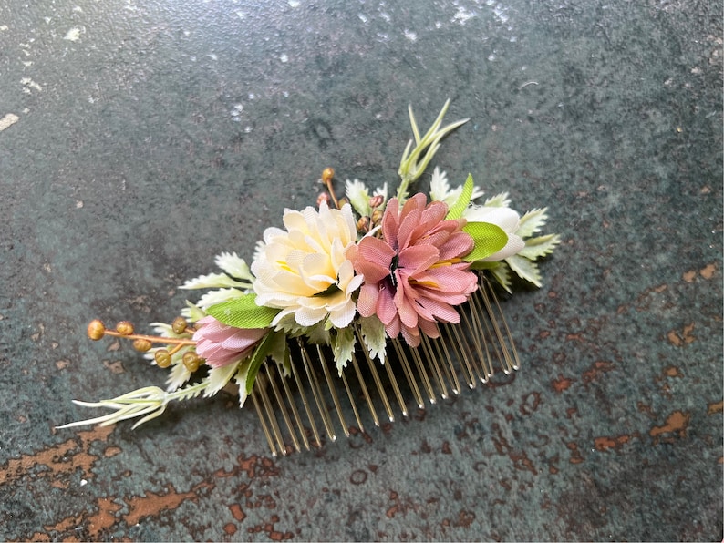 May include: A floral hair comb featuring a cluster of artificial flowers in shades of cream, pink, and mauve, accented with green leaves and sprigs. The flowers are attached to a gold-colored metal comb, suitable for weddings or special occasions.