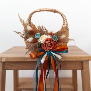 May include: Wicker basket decorated with dried flowers and ribbons. The arrangement includes orange, teal, and white ribbons, with a mix of beige, orange, and blue flowers. The basket sits on a wooden stool.