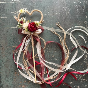 May include: A heart-shaped wedding wand with a twine frame, adorned with burgundy and cream-colored flowers and ribbons. The wand features a wooden stick handle and flowing ribbons in burgundy, cream, and white.