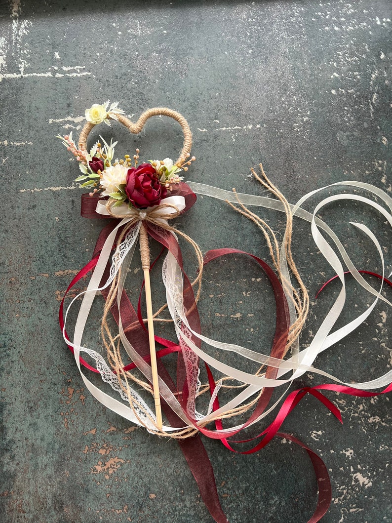 May include: A heart-shaped wedding wand with a twine frame, adorned with burgundy and cream-colored flowers and ribbons. The wand features a wooden stick handle and flowing ribbons in burgundy, cream, and white.