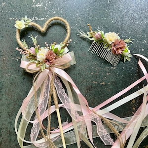 May include: A heart-shaped wand and a floral hair comb. The wand features a heart-shaped frame wrapped in twine, adorned with flowers, a bow, and ribbons. The comb has flowers and greenery. The color palette includes pink, white, and beige.
