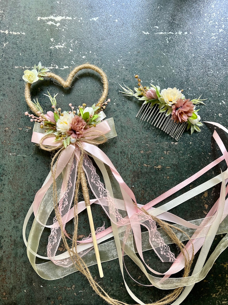 May include: A heart-shaped wand and a floral hair comb. The wand features a heart-shaped frame wrapped in twine, adorned with flowers, a bow, and ribbons. The comb has flowers and greenery. The color palette includes pink, white, and beige.