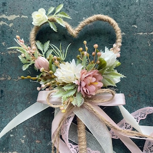 May include: A heart-shaped floral wand with a jute-wrapped frame. The wand features white, pink, and green artificial flowers, with a pink ribbon and lace bow. The handle is wrapped in jute, and pink ribbons cascade down.