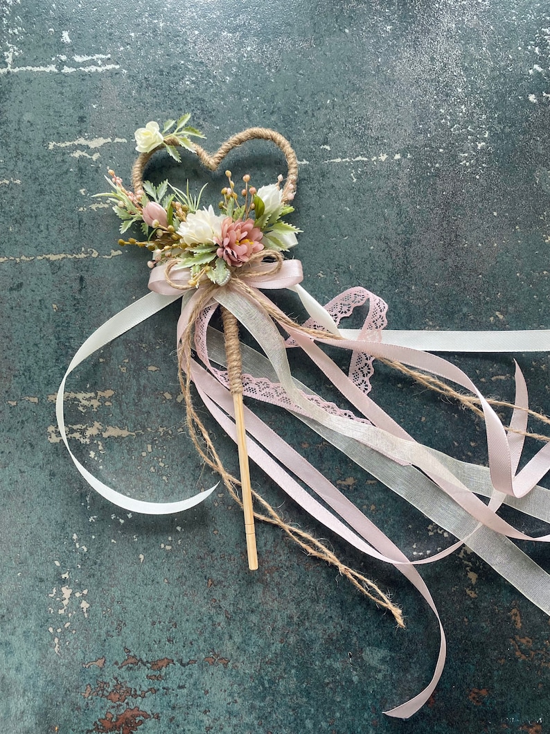 May include: A heart-shaped wedding wand with a floral arrangement of white and pink flowers, greenery, and a jute-wrapped handle. The wand features flowing ribbons in white, pink, and lace.