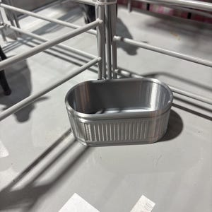 May include: A small, silver-colored metal trough with a ribbed exterior. The trough is oval-shaped and sits on a light gray surface. The background includes a white metal fence and a glimpse of a black animal.