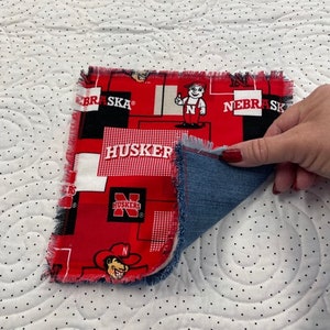 May include: A red and black fabric square with the words "Nebraska" and "Huskers" printed on it. The fabric is frayed around the edges and has a denim backing.