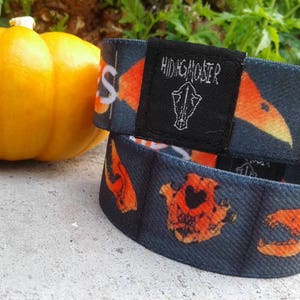 May include: Two black wristbands with orange and yellow designs. The top wristband has the text "HIDING MONSTER" on a black tag. The bottom wristband has a skull and crossbones design.
