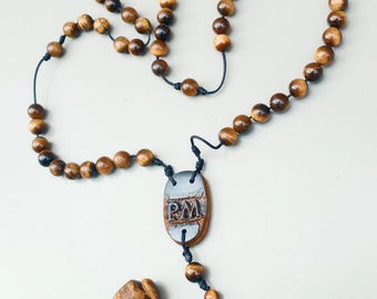 May include: A wooden rosary with brown beads and a dark cord. The rosary features a pendant with the letters "PM" and a carved wooden figure. The beads have a tiger eye appearance.