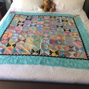 May include: A handmade quilt with a patchwork design. The quilt features a central section of colorful, patterned squares, bordered by a black dotted line and a turquoise floral fabric. A teddy bear sits on the bed.