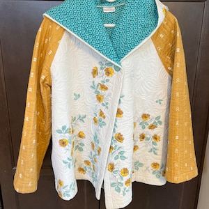 May include: A quilted jacket with a hood, featuring a white body with yellow floral embroidery. The sleeves are mustard yellow with a white arrow pattern. The hood lining is teal with a geometric design. The jacket is hanging on a white hanger.