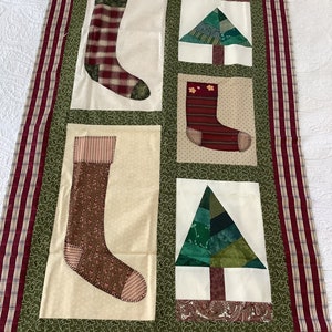 May include: A Christmas-themed quilt featuring four panels with appliqued stockings and Christmas trees. The stockings are in various patterns, including plaid and striped designs. The quilt has a green background and a red and white striped border.
