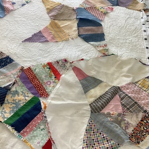 May include: A partially assembled quilt top featuring a starburst design. The quilt is composed of various fabric scraps in a range of colors and patterns, including floral, striped, and solid fabrics. The quilt is laid on a white, textured surface.