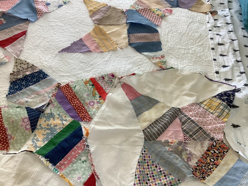 May include: A partially assembled quilt top featuring a starburst design. The quilt is composed of various fabric scraps in a range of colors and patterns, including floral, striped, and solid fabrics. The quilt is laid on a white, textured surface.