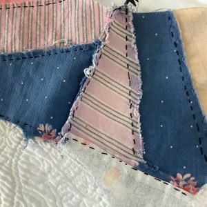 May include: Close-up of a patchwork textile featuring various fabric scraps. The design includes blue polka dots, pink stripes, and floral patterns, all stitched together with black thread. The background is a white, textured fabric.