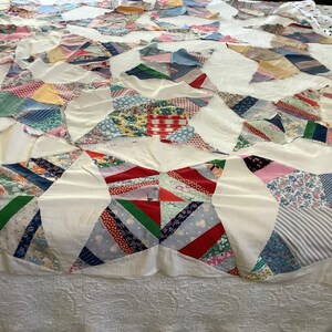 May include: A handmade quilt with a starburst pattern. The quilt features a patchwork design with various fabric patterns and colors, including red, blue, green, and floral prints. White fabric separates the starburst shapes. The quilt is laid out on a bed.