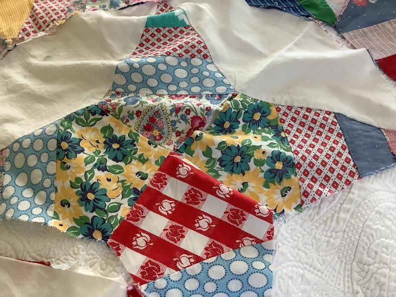 May include: Close-up of a handmade quilt featuring a starburst pattern. The quilt showcases a variety of fabric patterns, including floral, polka dots, and gingham in shades of blue, red, yellow, and white. The quilt is made of multiple fabric pieces.