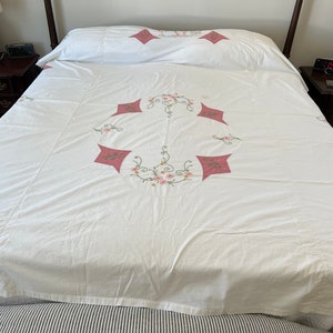 May include: White bedspread with a pink floral embroidery design. The embroidery features a central circle with pink flowers and green leaves, surrounded by four pink diamond shapes.