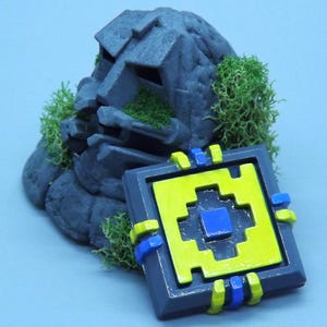 May include: A gray stone sculpture with green moss and a pixelated yellow, blue, and gray square. The square has a blue center and yellow accents. The sculpture resembles a face.