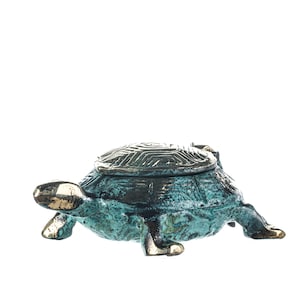 Könnte beinhalten: Bronze-farbene Schildkrötenfigur mit aufklappbarem Panzer. Der Körper der Schildkröte hat eine Grünspan-Oberfläche, während Kopf und Füße goldfarben sind. Der Panzer weist ein geometrisches Muster auf. Auf weißem Hintergrund.