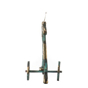 May include: A bronze-colored miniature chariot with two wheels and a vertical structure. The chariot has a weathered, aged appearance with hints of green and gold accents. The wheels are circular and the chariot is set against a white background.