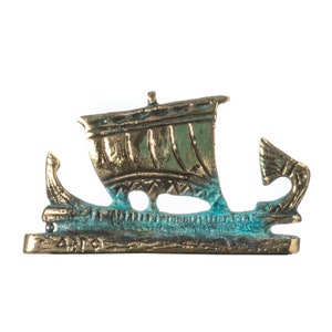 May include: A bronze-colored, stylized ship figurine with a detailed sail and decorative elements. The ship has a weathered, aged appearance with hints of blue-green patina. The base of the ship has Greek text.