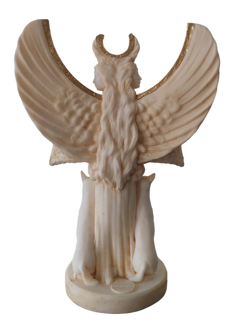 May include: White marble statue of a two-faced goddess with wings and a crescent moon on her head. The goddess is holding two white cats in her arms. The statue is on a round base with a gold inscription.