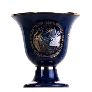 Puede incluir: Un jarrón de cerámica azul oscuro con borde y base dorados. El jarrón presenta un diseño circular con figuras y la palabra "GREECE" en oro. El diseño incluye un cisne y figuras de estilo clásico.