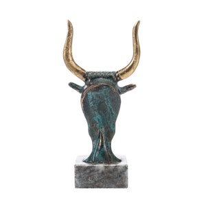 May include: A bronze bull head sculpture with gold horns on a marble base. The bull's head is facing forward with its horns pointing upwards.